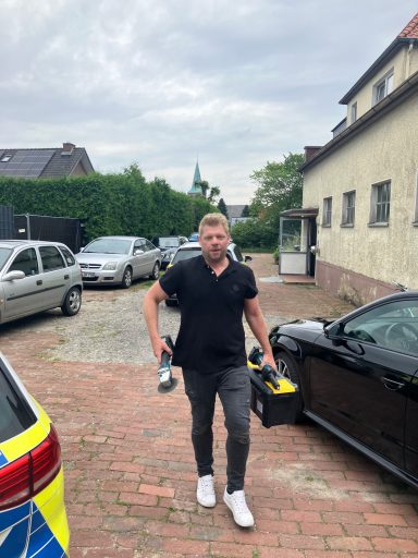 Mann in schwarzem Polo-Shirt mit Werkzeugkoffer läuft zwischen parkenden Autos.