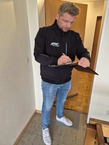 Mann in schwarzer Jacke notiert etwas auf einem Clipboard in einem Flur.