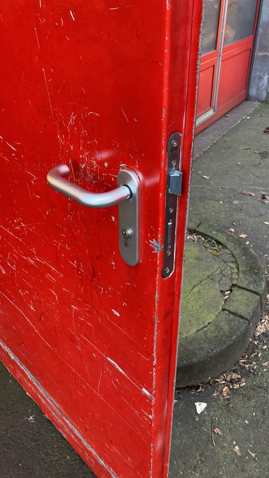 Rote Tür mit verkratzter Oberfläche und silbernem Türgriff, leicht geöffnet.