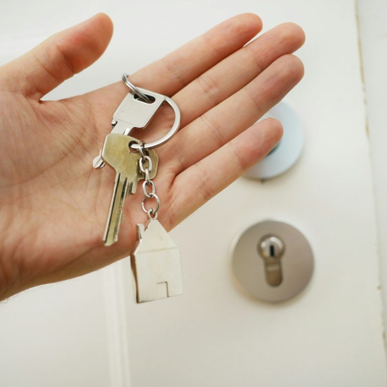 Ein Hand hält Schlüssel mit einem Schlüsselanhänger in Form eines Hauses vor einem Schloss.