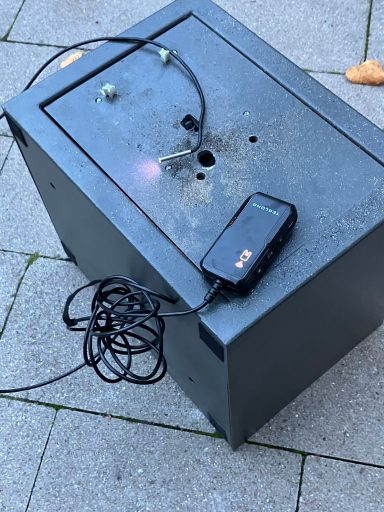Schwarzer Kasten mit Kabel und einer Box darauf, auf einem gepflasterten Boden.