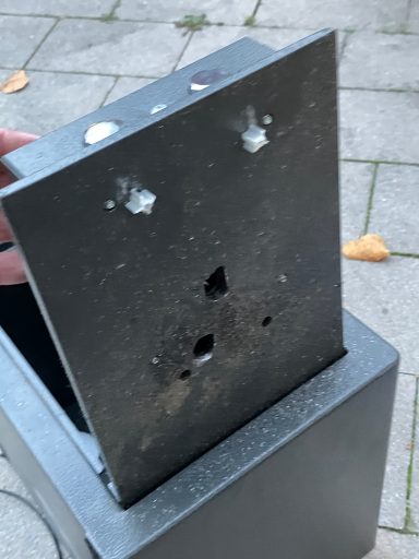 Oben auf einem grauen Metallkasten sind Schraubenlöcher und ein beschädigtes Metallstück sichtbar.
