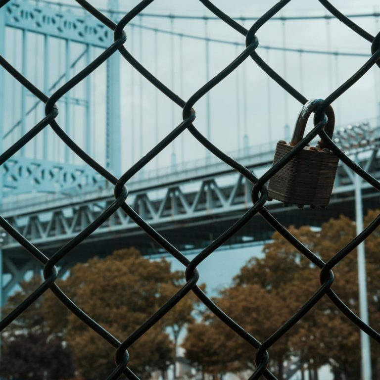 Ein dichter Zaun mit einem Schloss, im Hintergrund eine Brücke und herbstliche Bäume.