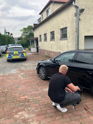 Mann kniet neben einem schwarzen Auto, Polizeiauto im Hintergrund auf einem gepflasterten Weg.