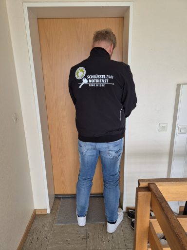 Person mit schwarzer Jacke steht vor einer geschlossenen Tür in einem Flur.